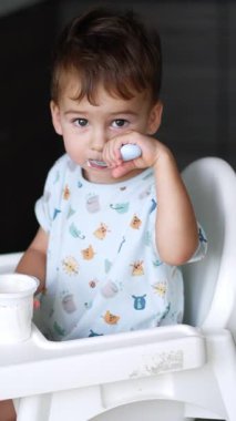 Sevimli gülümseyen erkek bebek yüksek masada oturuyor. Bebek kaşığı ağzında, diğer elinde yoğurt olan bir fincan tutuyor. Üst Manzara. Dikey video