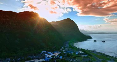 Güneş kayanın arkasından parlıyor. İnsansız hava aracı Pasifik Okyanusu kıyısındaki yerleşim alanından yükseliyor. Bulutlar gökyüzünde pembeye boyanmış. Hawaii, ABD.
