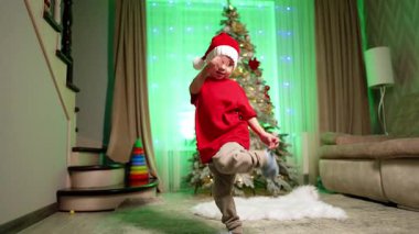 Noel Baba şapkalı aktif beyaz çocuk evdeki Noel ağacının yanında dans ediyor. Küçük çocuk tatilde eğleniyor..