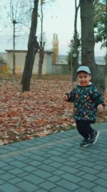 Parkta koşan, sıcak kıyafetler giyen neşeli bir oğlan çocuğu. Sonbahar günü yürüyüşe çıkmış küçük bir çocuk. Dikey video