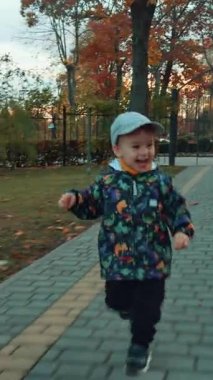 Parkta koşuşturan neşeli enerjik çocuk. Sonbaharda yürüyüş sırasında aktif zaman geçiren sevimli gülümseyen bebek. Dikey video
