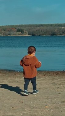 Kahverengi ceketli bir çocuğun nehre doğru yürüdüğünü gördüm. Bebek suya birkaç taş atar ve yenilerini aramak için döner. Dikey video