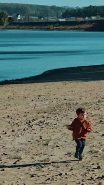 Nehir kıyısında koşan kahverengi ceketli küçük bir çocuk. Doğada suyun yanında yürüyen bir çocuk. Dikey video