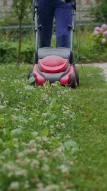 İnsan, yazın bahçedeki patikanın yakınındaki çim biçme makinesinin işini kontrol ediyor. Çim biçme makinesi sürekli olarak çimleri biçiyor. Dikey video.