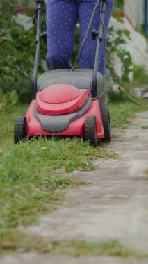 Kız yazın bahçede güllerle birlikte çiçek saksılarının etrafındaki çim biçme makinesiyle çim biçiyor. Çimleri biçmeye yakın çekim. Dikey video