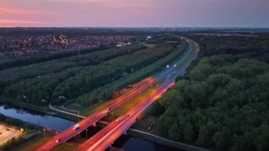 Kanal üzerindeki yol köprüleri gün batımında pembeye boyandı. Arkaplanda yemyeşil orman ve şehrin ışıkları var. Hava perspektifi.