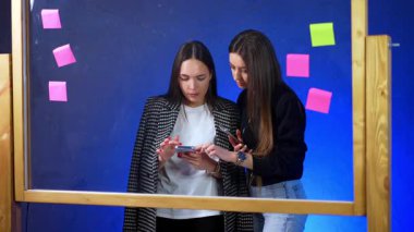 Beyaz kadınlar cam panonun önünde çıkartmalarla dikiliyorlar. Esmer kadınlar telefonları tutarlar ama ceketli kızın telefonuna odaklanırlar..