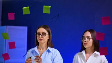 Gözlüklü ve gömlekli esmer kadın cam tahtasında bir kalemle ok çiziyor. Meslektaşım izliyor..