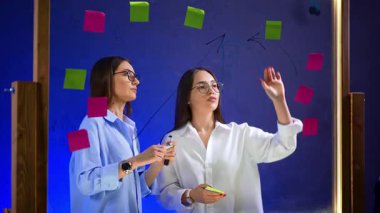 Ofisteki cam panoda meşgul bayanlar var. Meslektaşlar grafikler çizer ve camlara etiket yapıştırırlar. Fikirler ve planlar aranıyor.