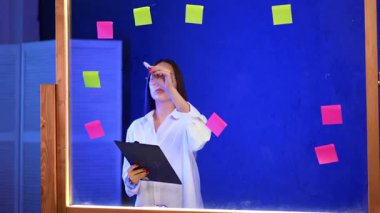 Uzun esmer saçlı odaklanmış genç bayan cam tahtaya yazı yazıyor. Ofiste elinde bir klip tahtası olan bir kadın..