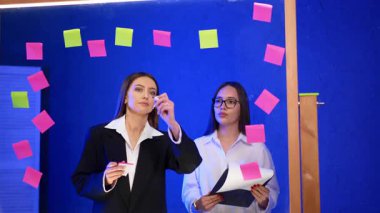 Modern ofis tezgahında çalışan bayanlar. Ceketli kadın bardağa yüzde numaraları yazıyor ve tişörtlü kadın elinde bir kupon tutuyor..