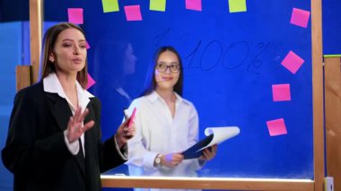 Siyah ceketli kadın profesyonel kameraya gülümsüyor ve camda yazan rakama işaret ediyor. Meslektaşım elinde bir klipsle cam tahtanın arkasında duruyor..