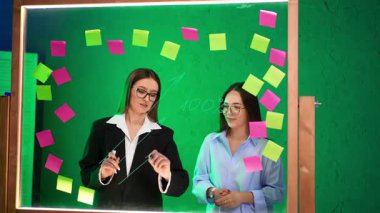 Ofiste fikirler üzerinde çalışan çekici kadınlar. Bayanlar cam tahta kullanır, üzerine yazı yazar ve çıkartmalara bakarlar. Beyin fırtınası konsepti.