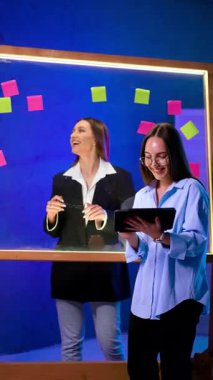 Pozitif kadın iş arkadaşları cam duvarın önünde duruyor. Hanımlar gülümser, konuşur, yapışkan notlara bakar. Fikir üretmek. Dikey video.