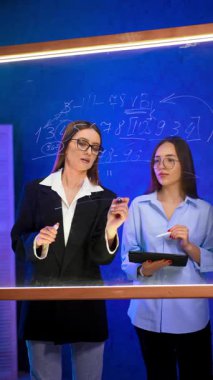 Çekici beyaz kadınlar cam duvarda yazan numaraları kontrol ediyorlar. Kadınlar iletişim kurar, panoya bakar ve birbirleriyle aynı fikirde olurlar. Dikey video.