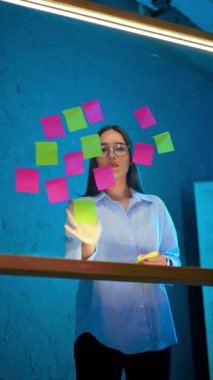 Beyaz gömlekli ve gözlüklü kadın cam tahtaya yapışkan notlar koydu. İş kadını planları hazırlıyor, cam duvarda notlar var. Dikey video.