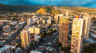 Batan güneşin son ışınları Honolulu 'nun yüksek binalarının tepelerini ve dağ yamacındaki evleri aydınlatıyor. Kara yağmurlu bulutlu manzara gökyüzünü kaplıyor. Hava perspektifi.