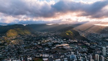 Batan güneş ışınları dağları aydınlatan gri bulutların arasından geçer. Honolulu, Hawaii, ABD 'nin yoğun şehir manzarasının üzerinde insansız hava aracı uçuşu..
