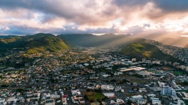 Batan güneş ışınları dağları aydınlatan gri bulutların arasından geçer. Honolulu, Hawaii, ABD 'nin yoğun şehir manzarasının üzerinde insansız hava aracı uçuşu..