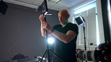 Fotoğraf stüdyosunda fotoğraf düzenleme ekipmanları. Sakallı kameraman mercekle tripodu ayarlıyor.