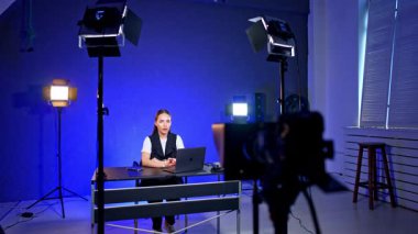 Beyaz bir kız stüdyoda dizüstü bilgisayarı ve telefonları açık bir şekilde konuşuyor. Fotoğrafçı masaya gelip bir şeyler alıyor..