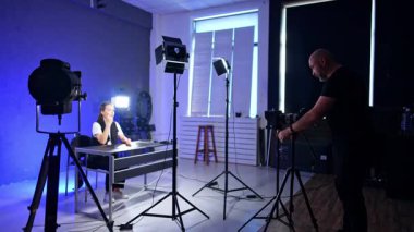 Çekimlerin yapıldığı modern bir fotoğraf stüdyosunun içinde. Esmer kız masada oturuyor. Kameraman gerekli ekipmanları ayarlar.