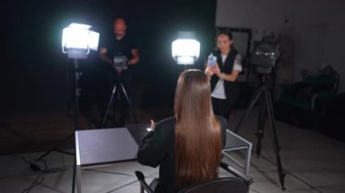 Uzun düz, esmer saçlı bir bayanın arkadan görünüşü masada oturup kameranın önünde konuşuyor ve el kol hareketi yapıyor. Soffit ışıkları muhabirin üzerinde. Kameraman ve kız arka planda telefonuyla çekim yapıyor..