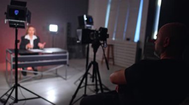 Kendinden emin, rahat erkek fotoğrafçı kameranın arkasında oturuyor. Siyah ceketli kadın masada oturuyor ve blog adına konuşuyor..