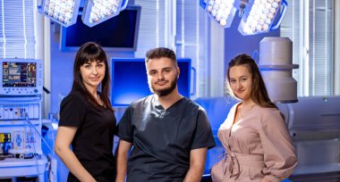 Tıbbi ekip ameliyathanede poz veriyor. Üç sağlık uzmanı, iyi donanımlı bir cerrahi ortamda, takım çalışması sergileyerek güvenle duruyorlar..