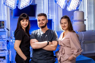 Tıbbi ekip ameliyathanede poz veriyor. Üç tıp uzmanı parlak bir cerrahi odada durup vardiyaları sırasında yoldaşlık ve güven gösteriyorlar..