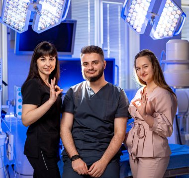 Modern bir klinikteki doktorlar. Üç sağlık çalışanı, iyi aydınlatılmış bir klinikte, önlükleri ve kıyafetleriyle güvenle sırıtarak bir arada duruyorlar..