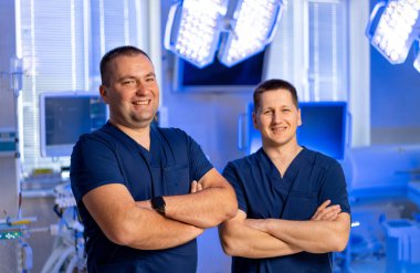 Hastanede gülümseyen tıbbi personel. İki erkek sağlık çalışanı yüksek teknolojili bir hastane odasında kollarını kavuşturarak güvenle duruyorlar..