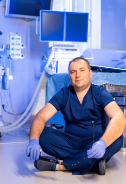 Cerrahlar ameliyat odasında. Mavi önlüklü bir cerrah elinde gözlüklerle hastane zemininde dinleniyor..