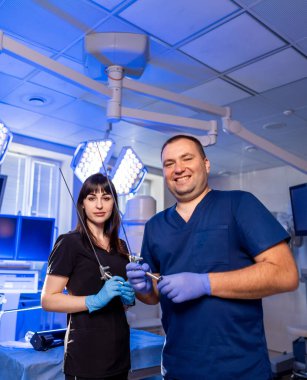 Klinik tıbbi personeli. İki sağlık çalışanı, modern, iyi ışıklandırılmış bir tıbbi ortamda cerrahi aletleri tutarken gülümsüyor.