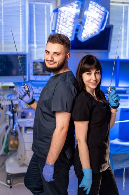 Cerrahi ekip ameliyat önlüğü içinde. İki sağlık uzmanı, araçlarını ve takım çalışmalarını vurgulayarak, parlak bir cerrahi ortamda güven içinde poz veriyorlar..