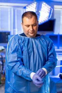 Cerrah ameliyata hazırlanıyor. Bir tıp uzmanı gündüz vakti ameliyat odasında mavi bir ameliyat elbisesi giyerken eldiven takıyor..