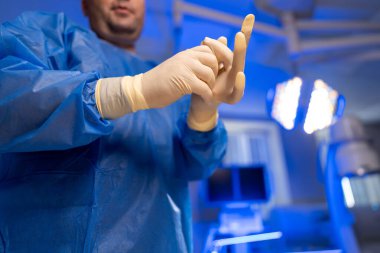 Cerrah ameliyata hazırlanıyor. Bir sağlık çalışanı cerrahi ortamda eldiven giyer, parlak ve steril bir odada ameliyata hazırlanır..