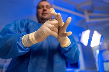 Cerrah ameliyata hazırlanıyor. Bir tıp uzmanı ameliyattan önce mavi ışıkla dolu bir ameliyathanede ameliyat eldiveni takıyor..