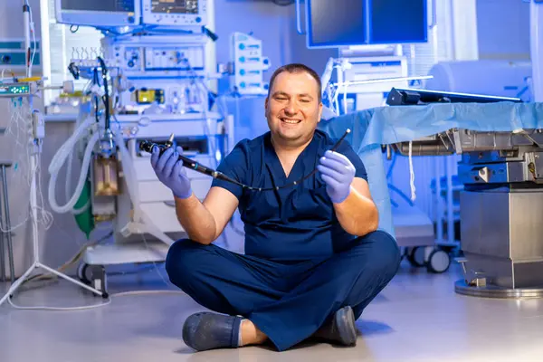 Doktor cerrahi alet gösteriyor. Bir sağlık çalışanı ameliyathanenin zemininde oturuyor, ameliyat aletini tutarken gülümsüyor..