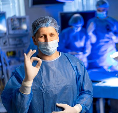 Ameliyathanede ameliyat ekibi var. Mavi önlüklü bir cerrah iyi aydınlatılmış bir ameliyathanede ameliyat yaparken iyi bir jest yapar..