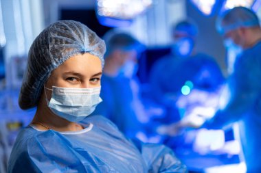 Ameliyathanede ameliyat ekibi var. Önlüklü ve maskeli bir sağlık uzmanı parlak bir ameliyathanede cerrahi müdahaleyi izler..