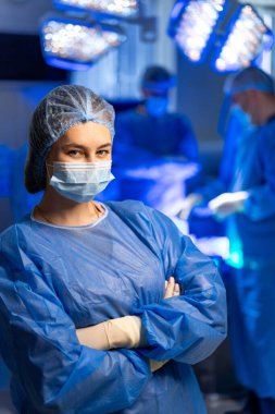 Cerrahi tıp uzmanı. Önlük ve maske takan kendine güvenen bir tıp uzmanı cerrahi düzende çalışan meslektaşları olarak hazır bekliyor..