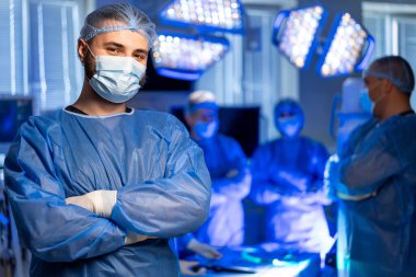 Cerrah ameliyathanede hazırlanıyor. Bir cerrah, parlak bir ameliyathane ortamında cerrahi bir ekibin önünde tamamen önlük ve maske takarak güvenle durur..