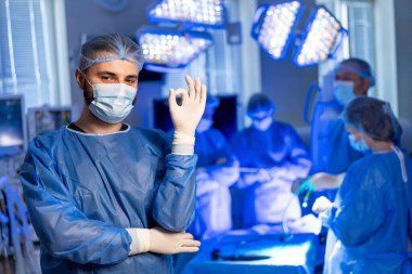 Ameliyat için ameliyat önlüğü giymiş bir cerrah hazır. Önlük giymiş bir cerrah, iyi ışıklandırılmış bir ameliyathanede bir ekiple ameliyata hazırlanırken kendinden emin bir şekilde hareket eder..