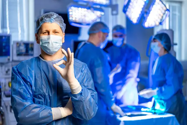 Ameliyathanede cerrahın parmağı var. Mavi önlüklü ve maskeli bir cerrah, meslektaşları ameliyat ederken, parlak cerrahi ışıklarla aydınlatılan, kendinden emin bir şekilde el kol hareketi yapar..