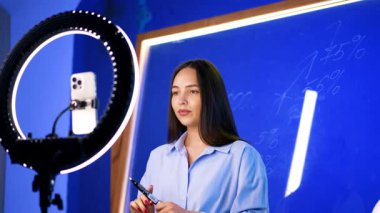 Uzun esmer saçlı, gömlek giyen bir kadın telefonuna yüzük lambasıyla bakıyor. Kadın cam duvarın önünde kamerayla konuşuyor. Günlük konsepti.