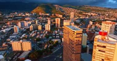 Parlak turuncu ışık binaların cephelerine yansıyor. Karanlık dramatik bulutlar arka planda dağlarda yuvarlanıyor. Honolulu, Hawaii, ABD.