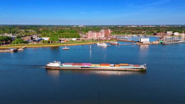Merkemeer Gölü 'nün su manzarasında ağır konteynırlarla dolu bir mavna hareket ediyor. Hollanda, Lelystad liman ve şehir manzarası arka planda. Hava perspektifi.
