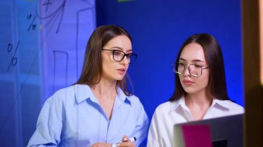 Gözlüklü ve beyaz gömlekli iki güzel beyaz kadın cam duvarda duruyor. Kadınlar konuşur, tartışır, plan yapar ve dizüstü bilgisayara bakar..