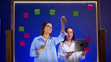 Gözlüklü esmer ve beyaz gömlekli cam tahtaya grafikler çiziyor. Yanında duran kız dizüstü bilgisayar tutuyor. İş stratejisi ve planlama.